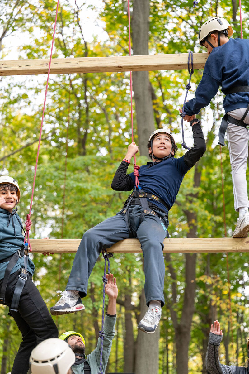 The image shows people on a ropes course. Several individuals are navigating the course, wearing helmets and safety harnesses. They are suspended in the air, with wooden beams and ropes supporting their progress. The background features lush green trees, suggesting an outdoor adventure setting.